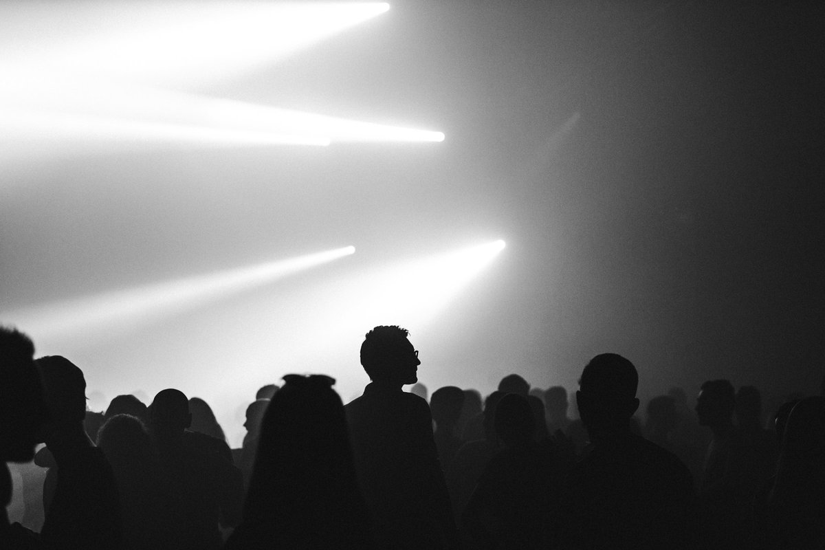 A black-and-white crowd in silhouette under bright beams of light cutting through haze, one man in glasses standing slightly above the heads around him