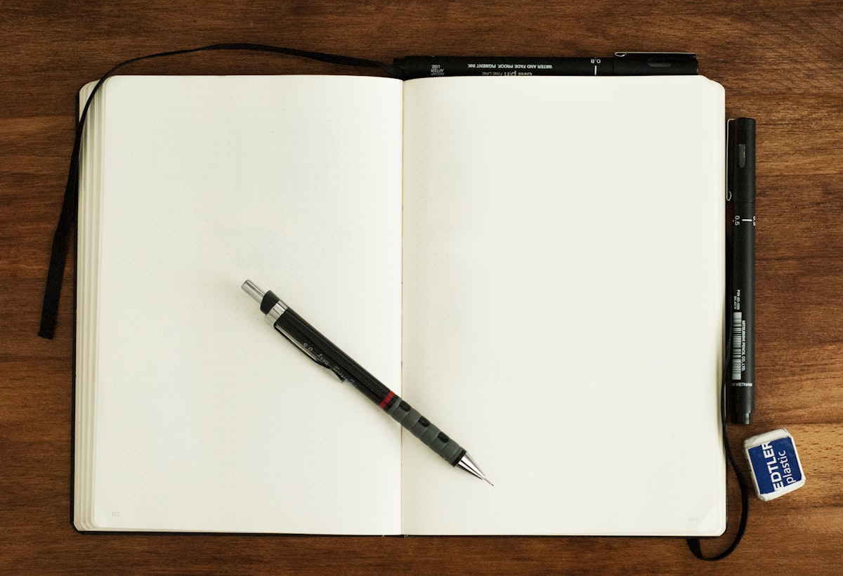 An open, blank sketchbook with drawing pens resting on it on a wooden desk