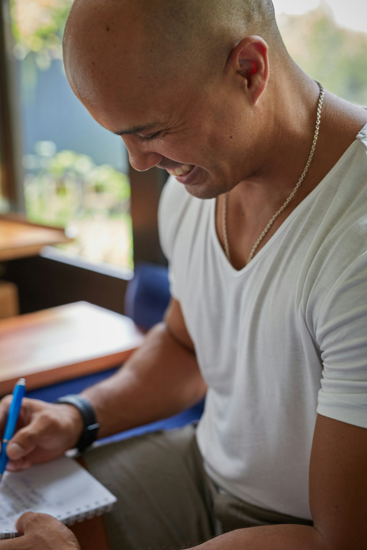 A man smiling broadly while writing a letter with a pen at a desk