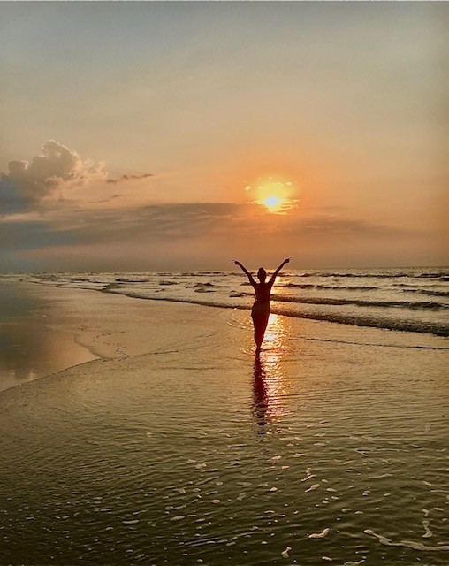 A silhouette with arms raised to a rising sun over the ocean at low tide