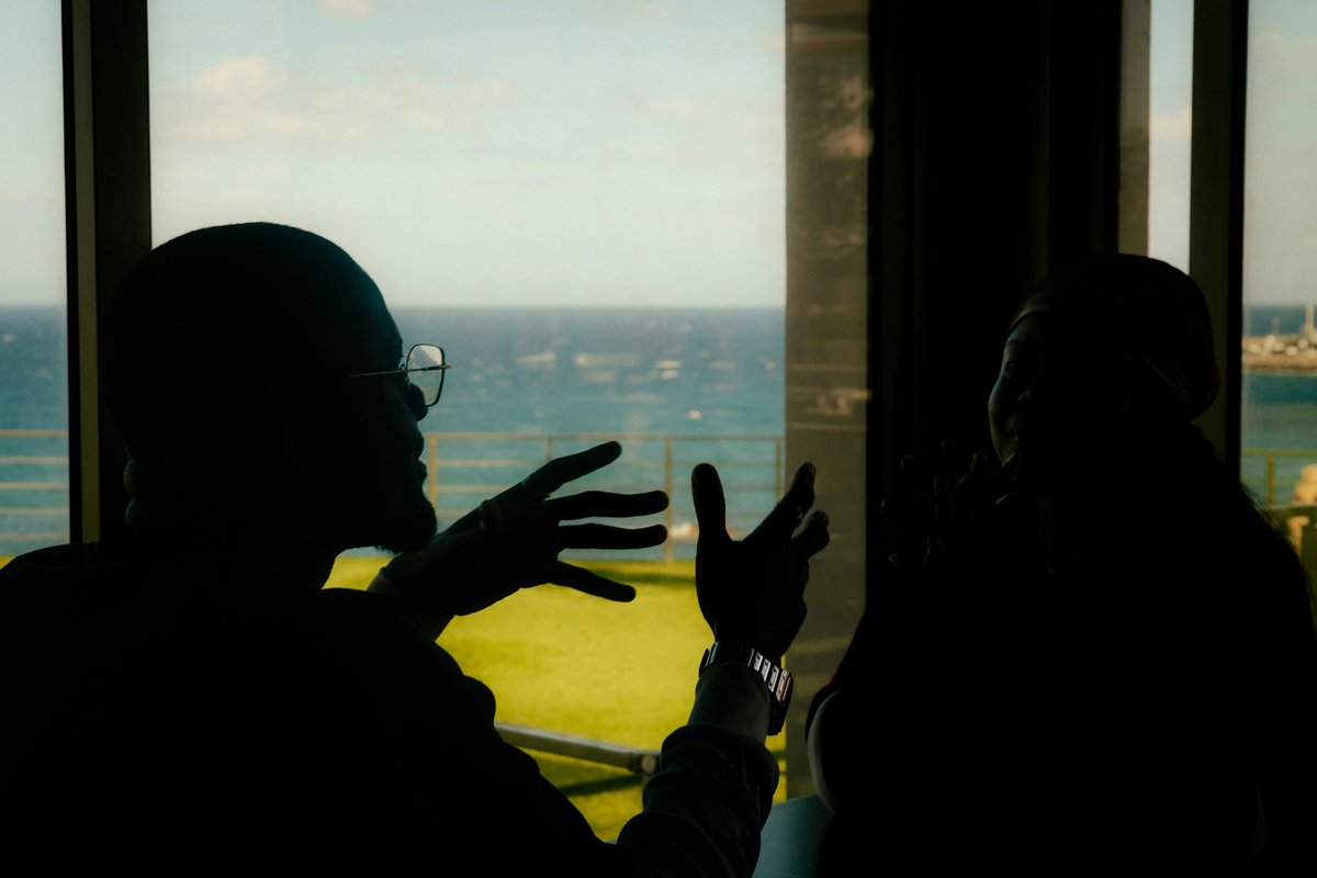 Two people in silhouette talking across a table by a tall window, one with glasses gesturing with both hands, blue sea and green lawn beyond the glass