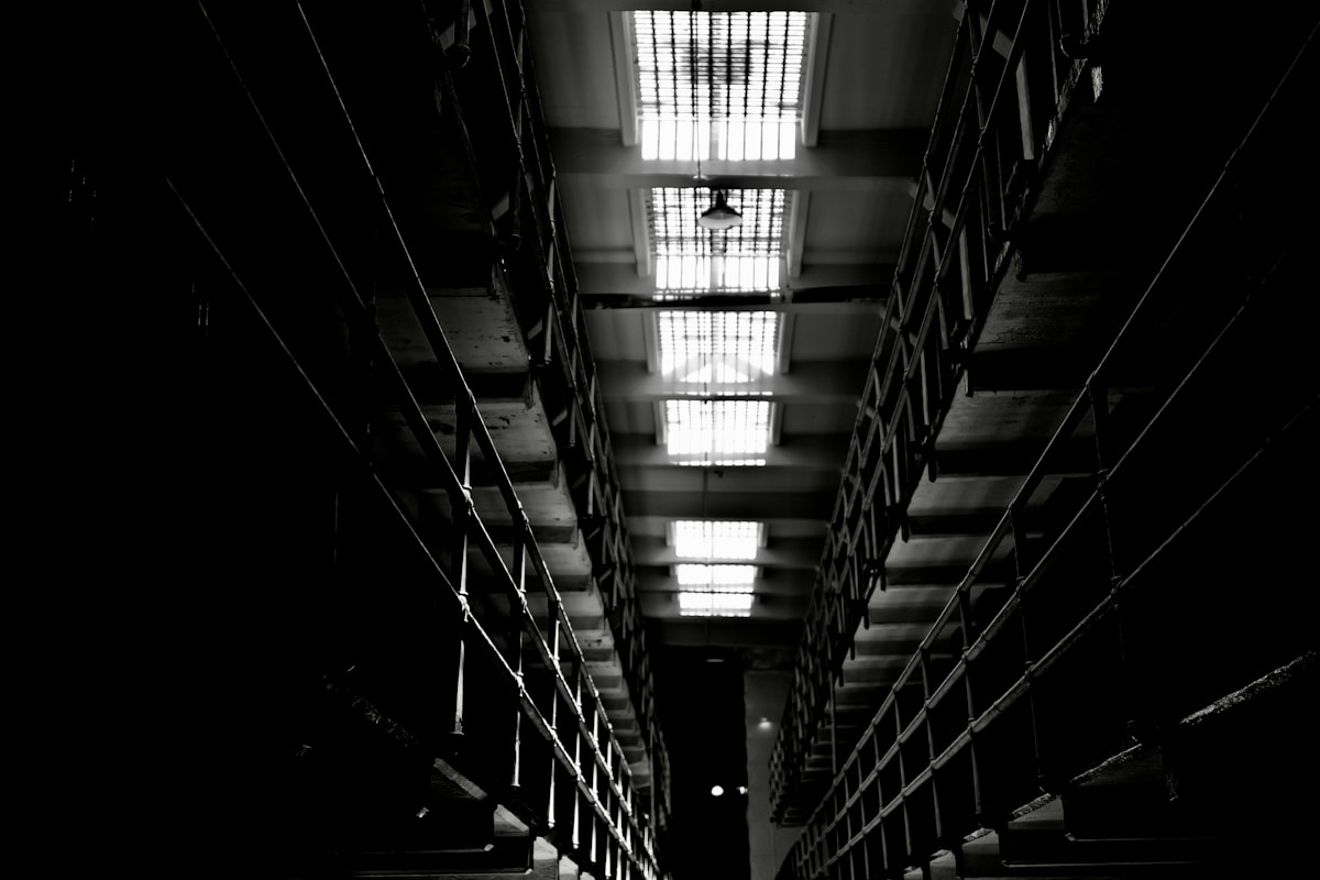A dark prison cell block photographed down its length, tiers of barred cell fronts receding into shadow beneath a row of bright barred skylights
