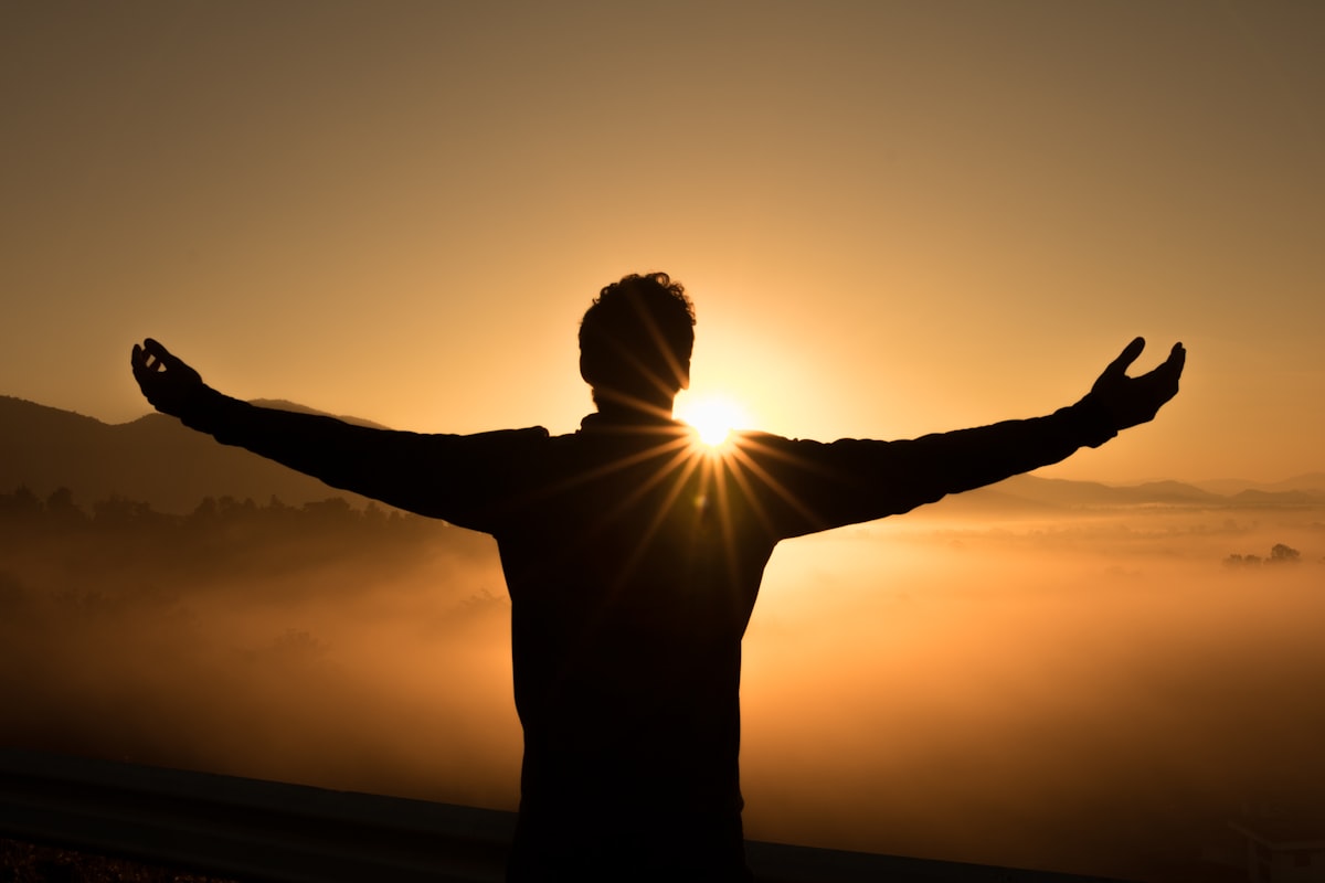 A lone person silhouetted against the sunrise with arms outstretched above a layer of mist