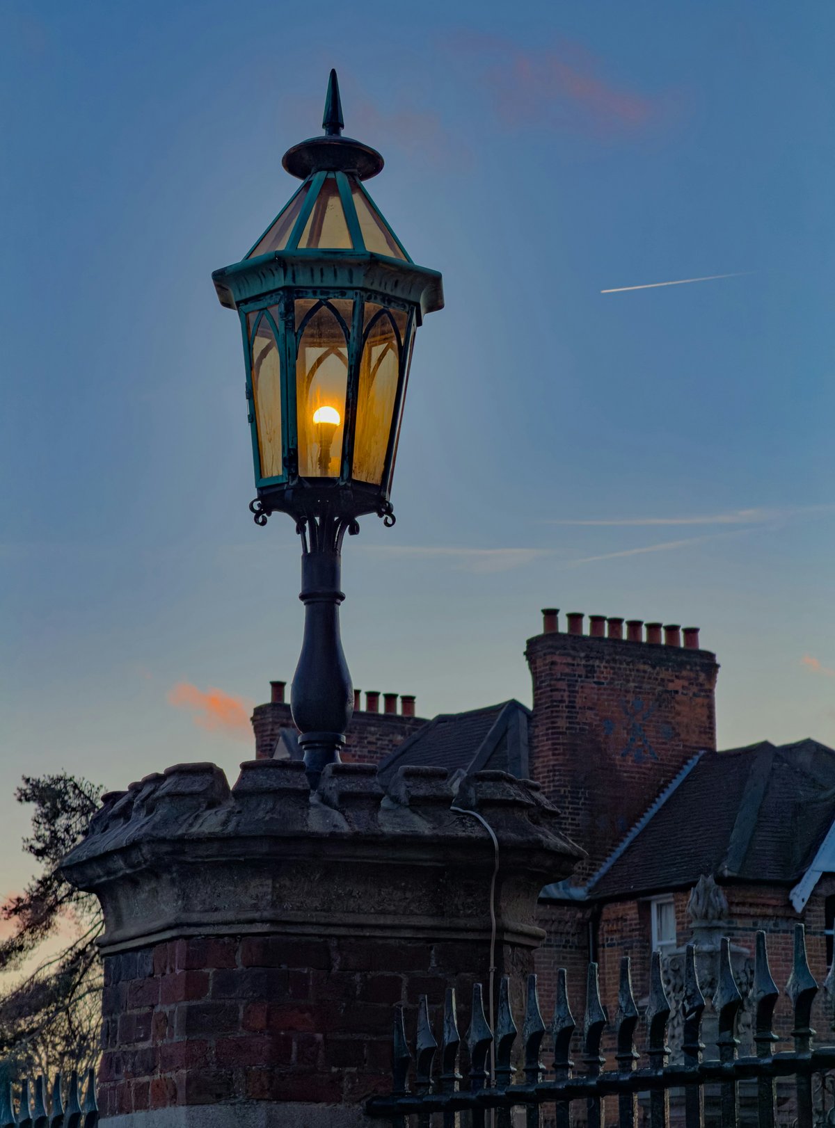 An ornate Victorian gas lantern burning amber against a deep blue twilight sky, mounted on a brick pier above iron railings, with tiled rooftops and chimney pots behind it