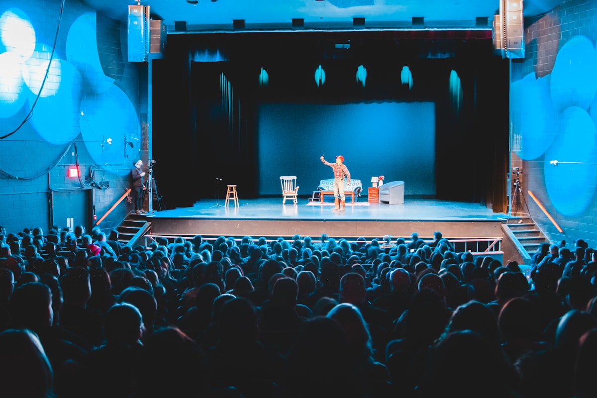 An empty comedy club stage with a single microphone stand under a spotlight, audience seats dark
