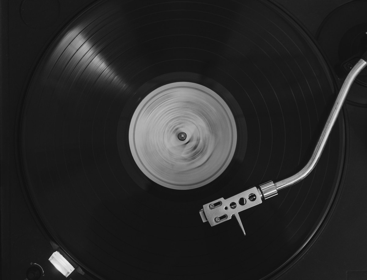 A black-and-white overhead view of a vinyl record spinning on a turntable, the label blurred by motion and the tonearm resting in the groove