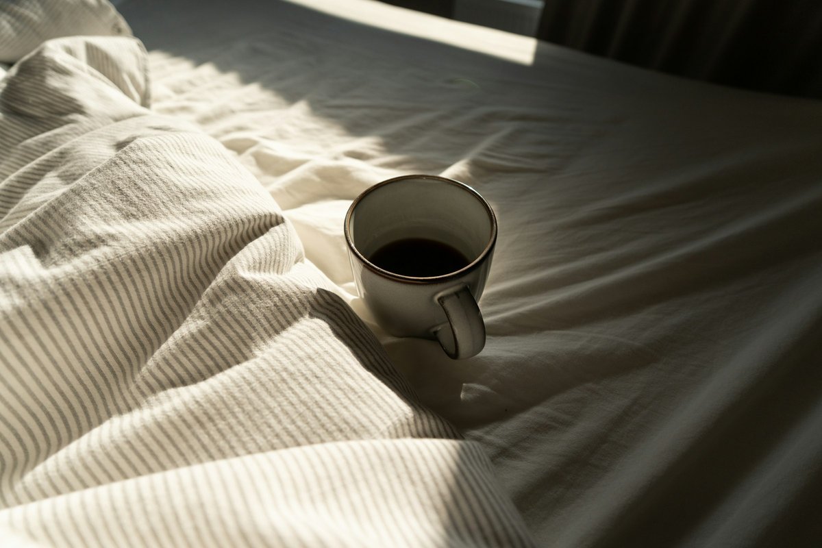 A cup of black coffee resting on rumpled white and grey striped bedding, a band of sunlight falling across the sheets
