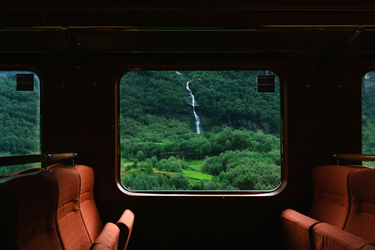 The inside of a dim train carriage with empty orange seats, its large window framing a green forested valley and a tall white waterfall